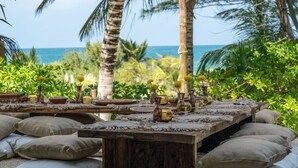 A wooden table set with pottery and cushions, surrounded by lush greenery and a view of the ocean. - Casa Maya Kaan (Tulum)