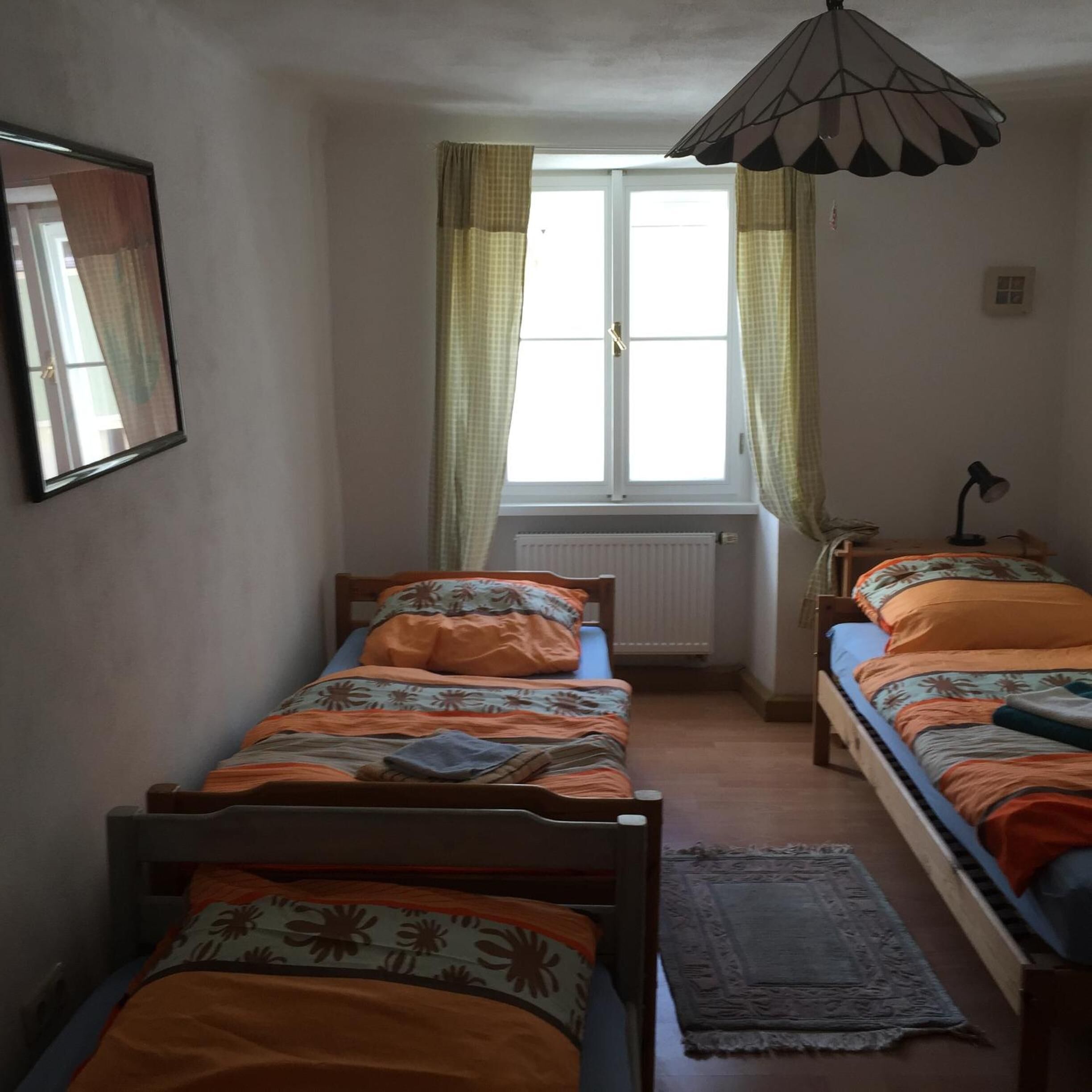 A dormitory room with two single beds, a window with curtains, a radiator, a desk with a lamp, and a mirror on the wall.