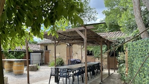 Un patio al aire libre con una pérgola de madera, una mesa larga y sillas. Se observan barriles y el tronco de un árbol en primer plano.