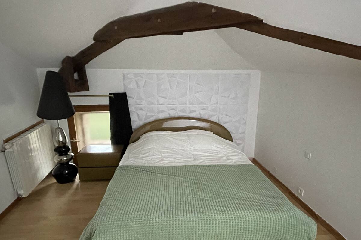 A bedroom with a sloped ceiling, a bed with a green blanket, a bedside lamp, and a radiator.