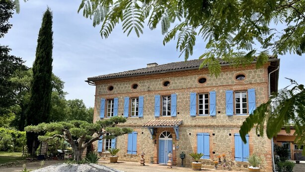 Un edificio de piedra con contraventanas azules, una puerta central y un patio de grava.