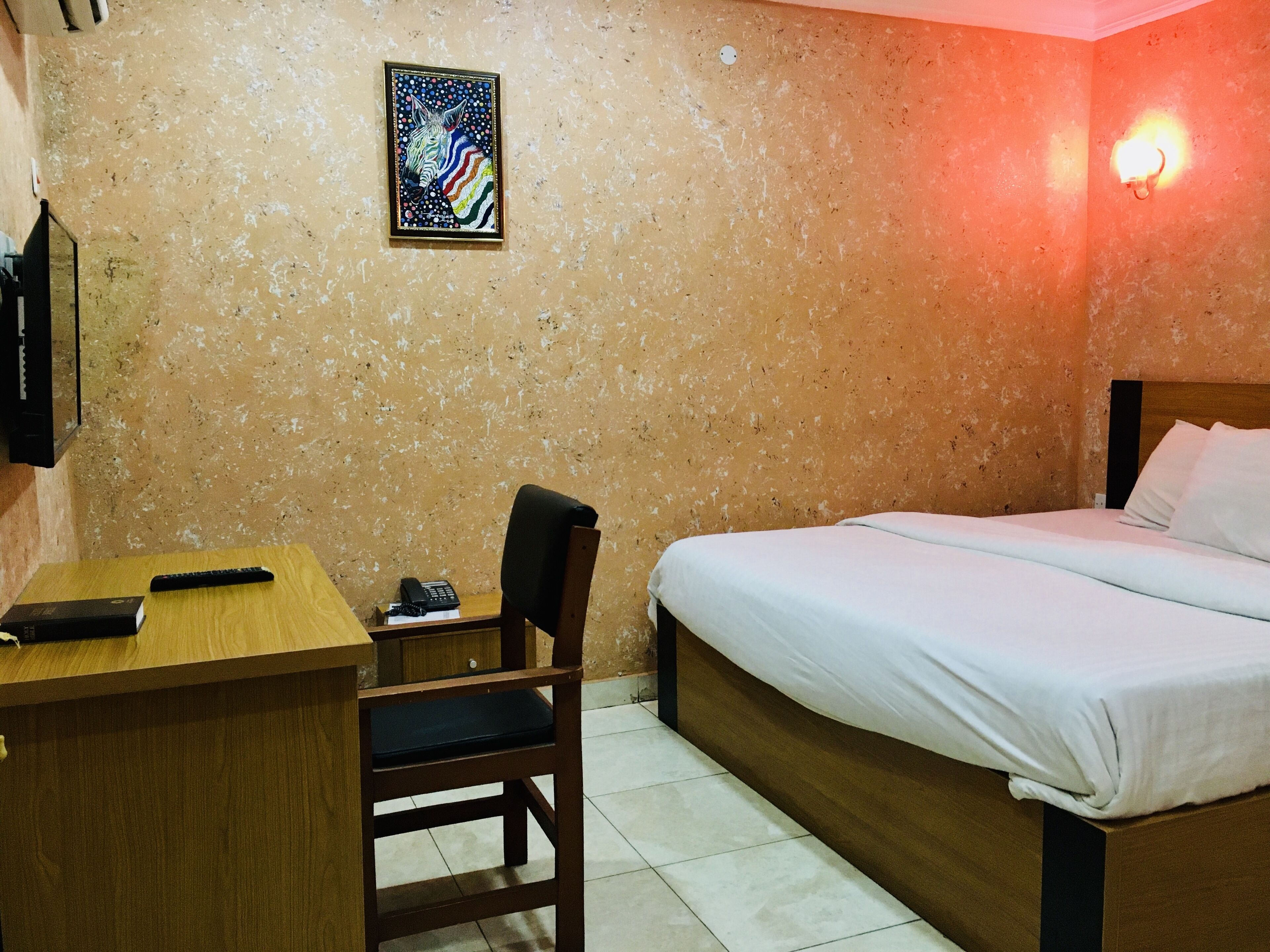 A bedroom with a bed, a desk, a chair, a television, and a wall-mounted light fixture.