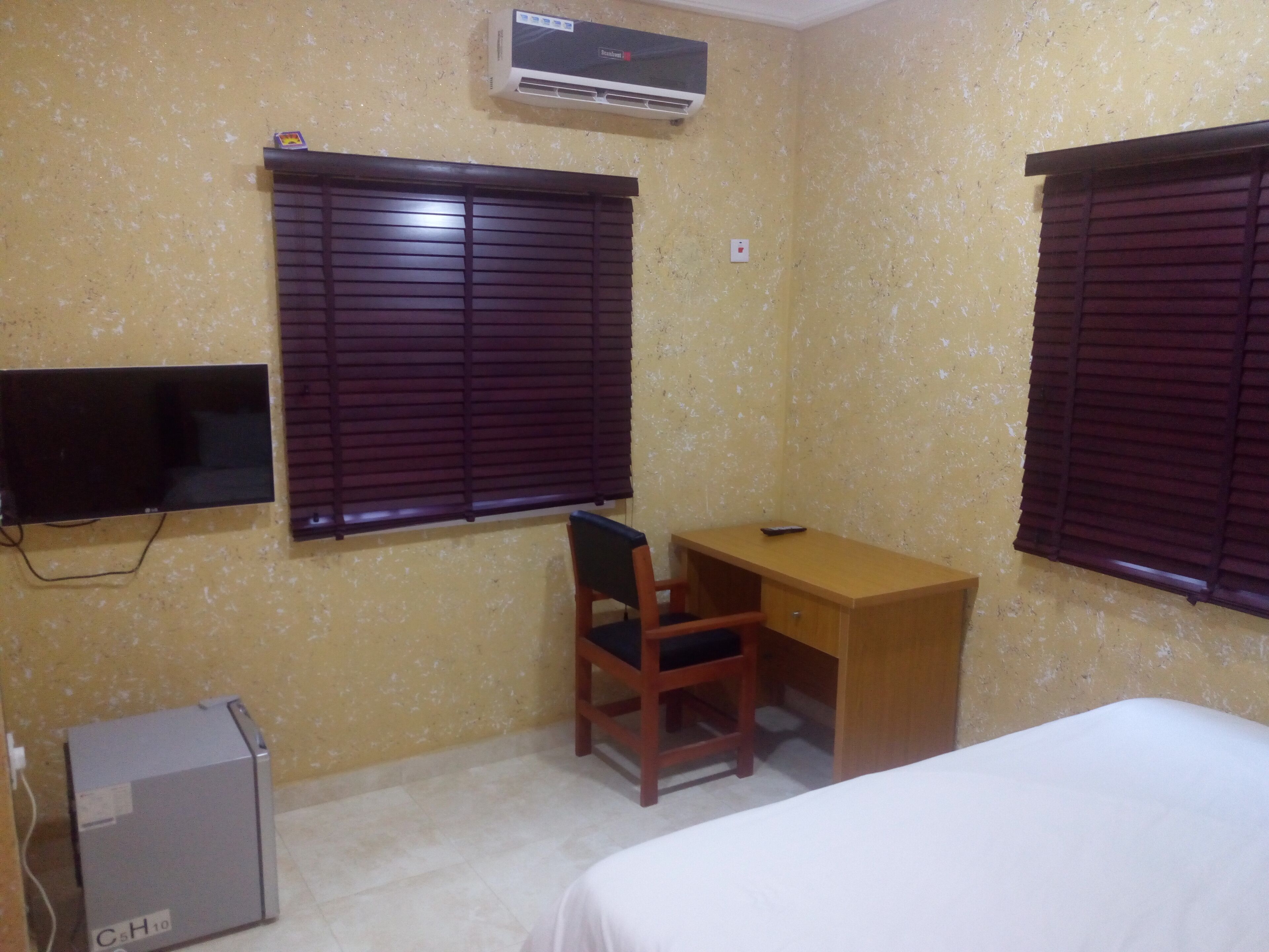 A small, simple room with two windows, a desk, a chair, a bed, and an air conditioner.