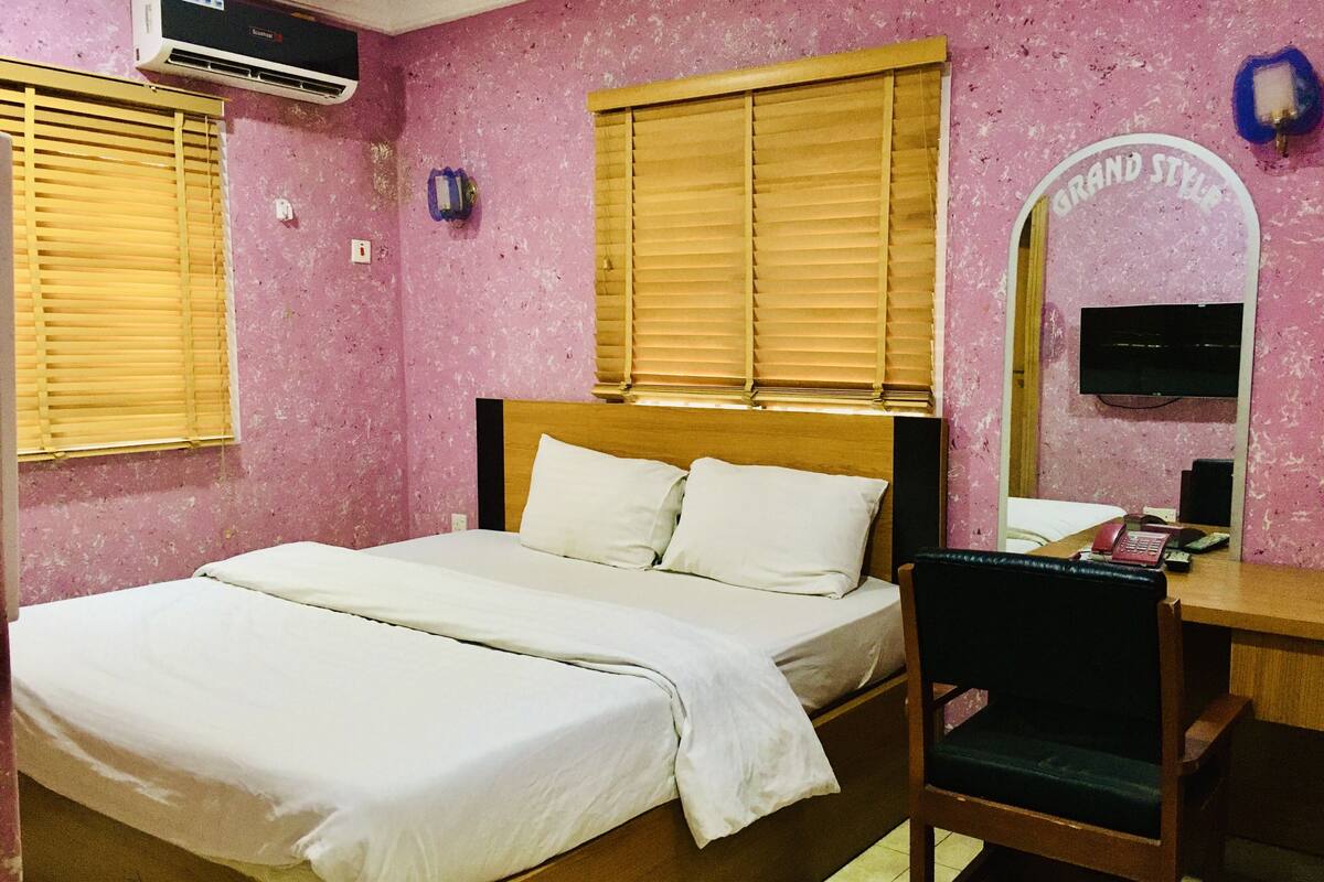 A bedroom with a bed, wooden headboard, two windows with blinds, an air conditioner, a desk with a chair, and a mirror.