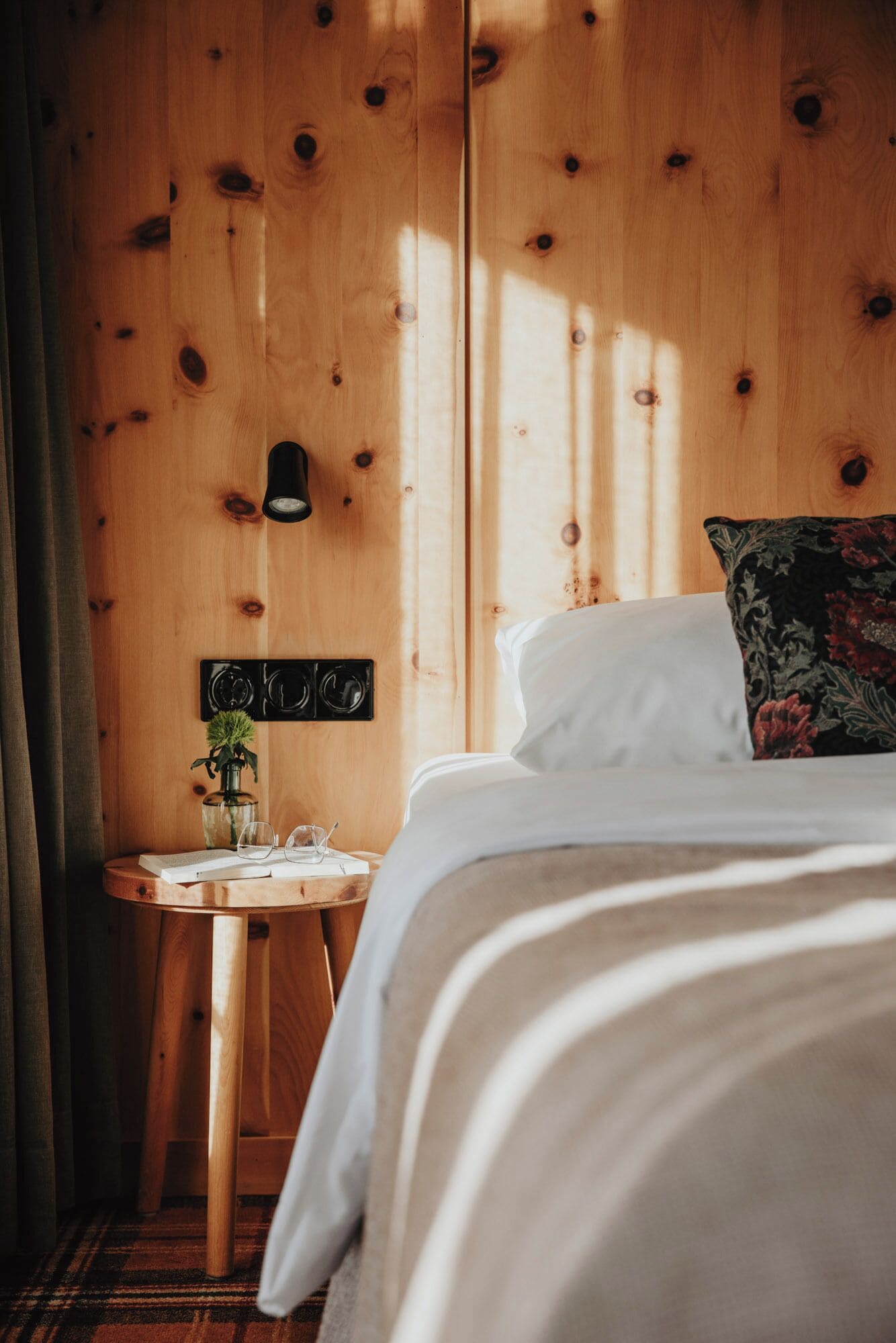 Una habitación de madera con una cama, una mesita con una planta y una lámpara fijada a la pared.