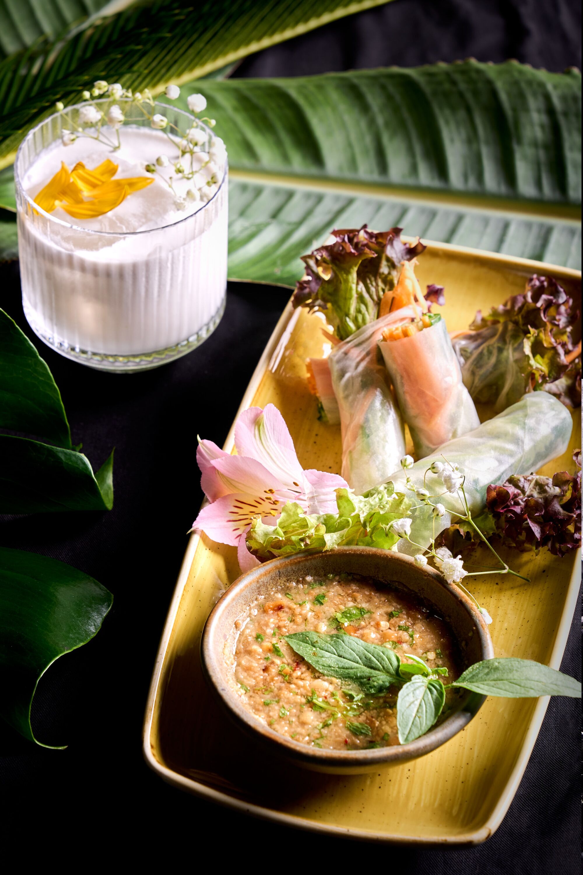 A tray with spring rolls, a bowl of dipping sauce, and a glass of white beverage with a flower garnish.