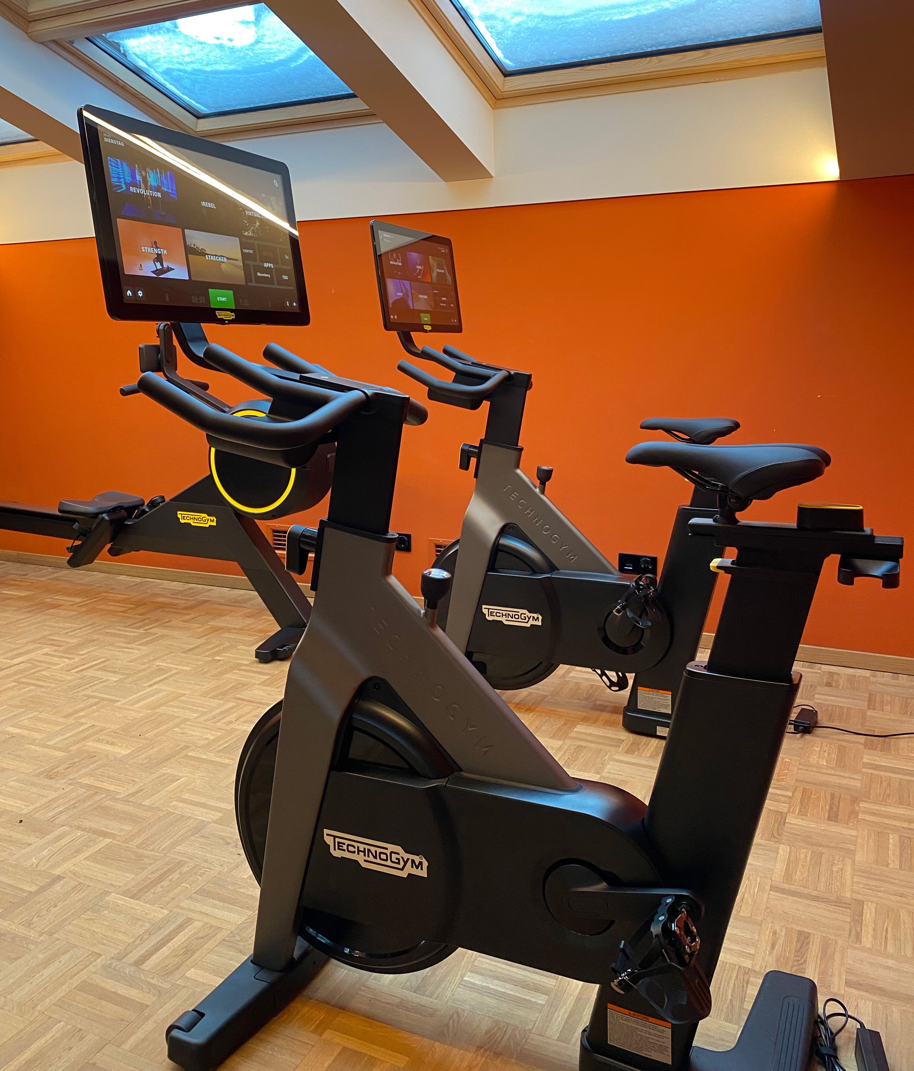 A gym with two Technogym stationary bikes, a digital display, and an orange wall.