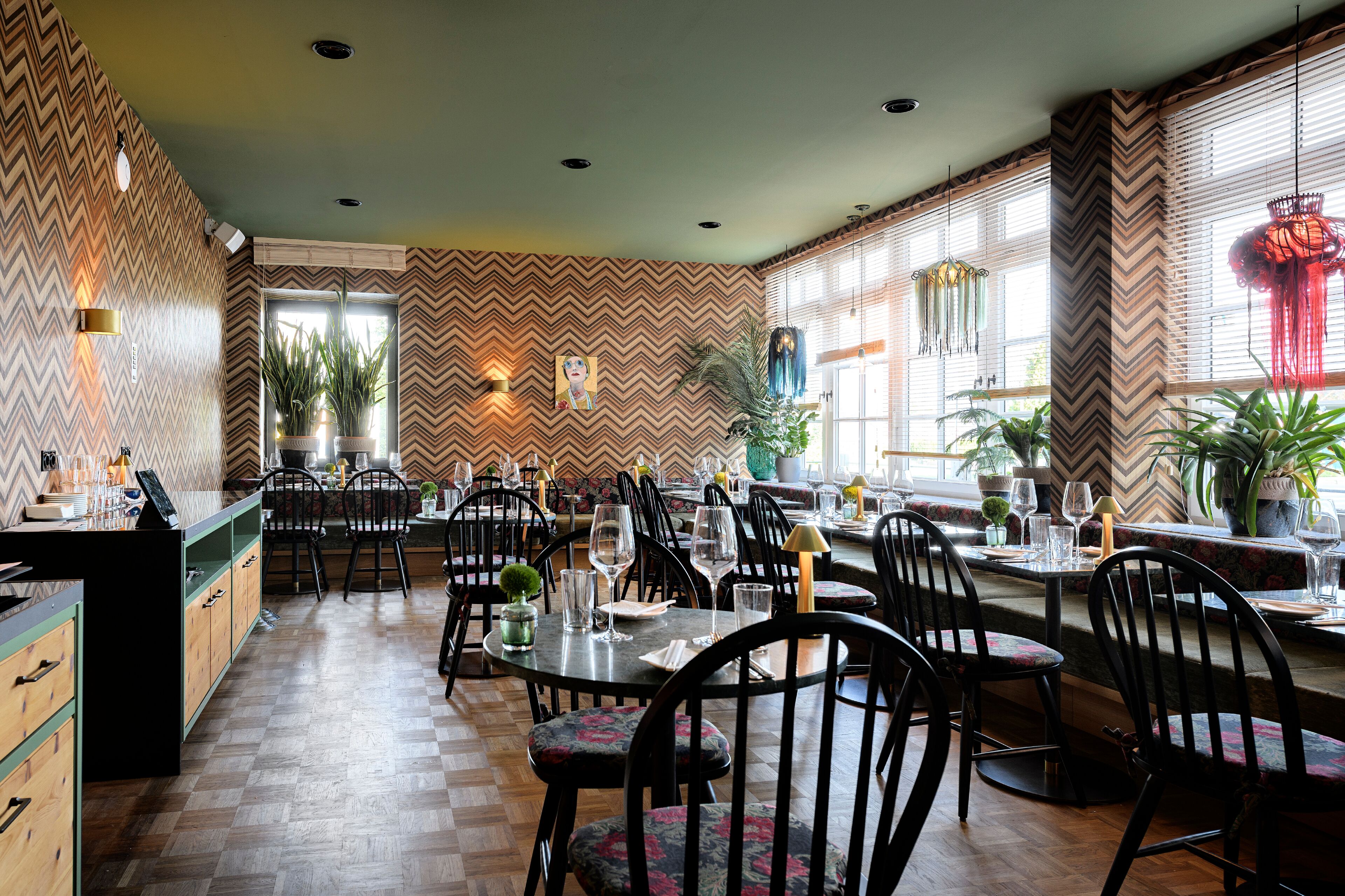 Um restaurante com parede em padrão geométrico em chevron, piso de madeira e cadeiras pretas com estofamento floral.