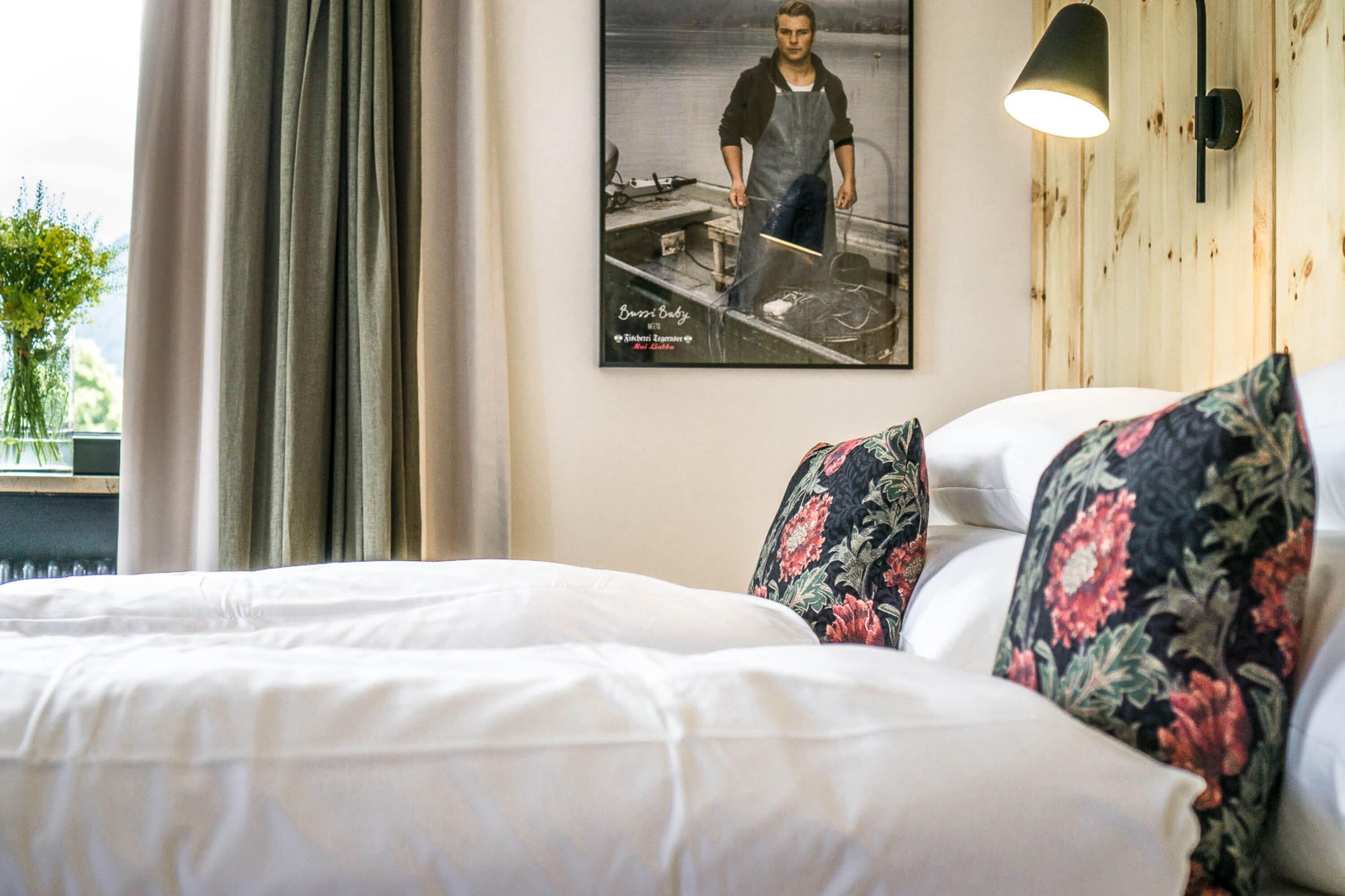 Una habitación de hotel con una cama, almohadillas estampadas, una lámpara de pared, un cartel y una ventana con cortinas.