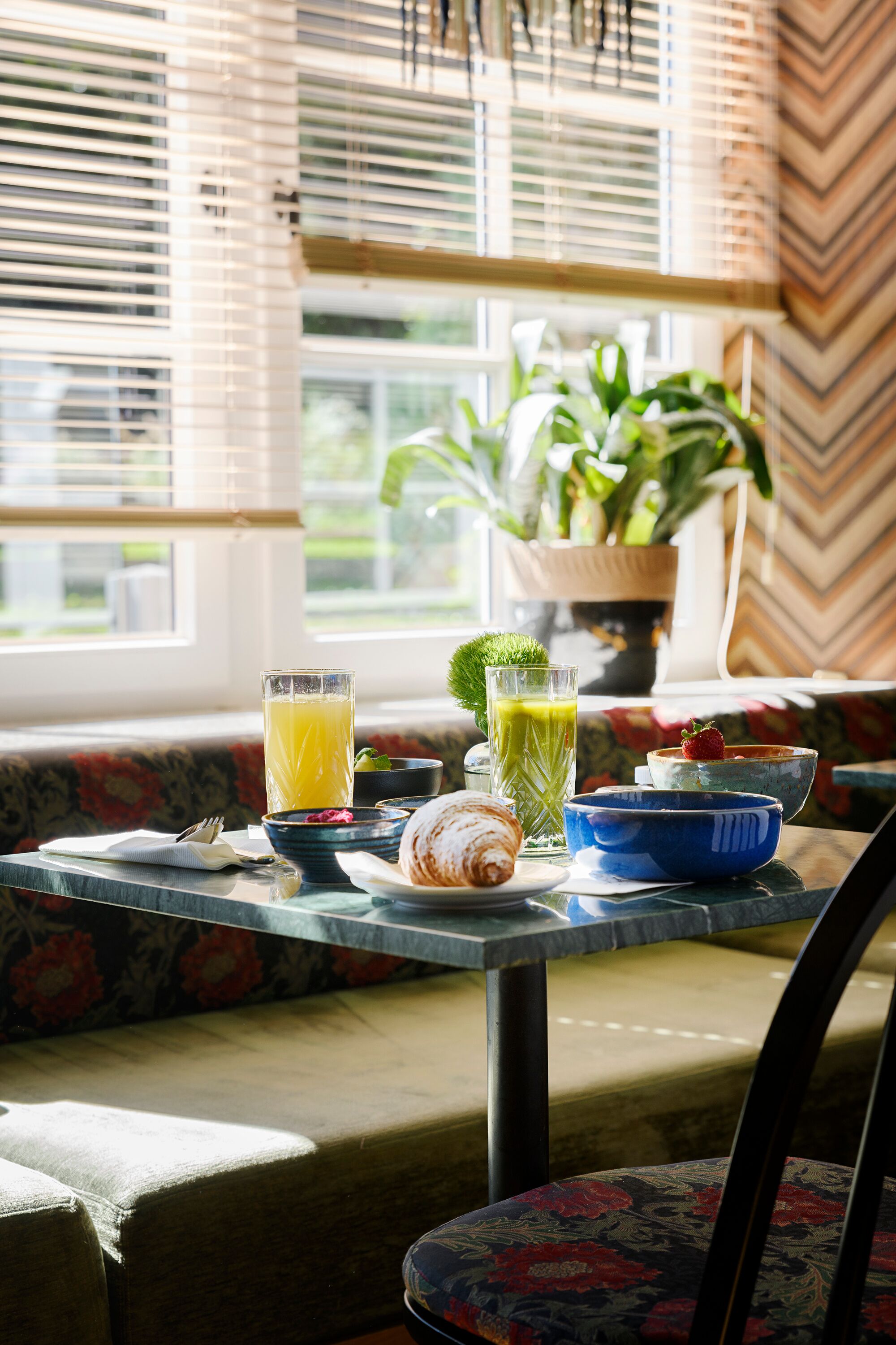 Una mesa de desayuno con un croissant, un vaso de jugo de naranja, un vaso de jugo verde y un tazón de fresas.
