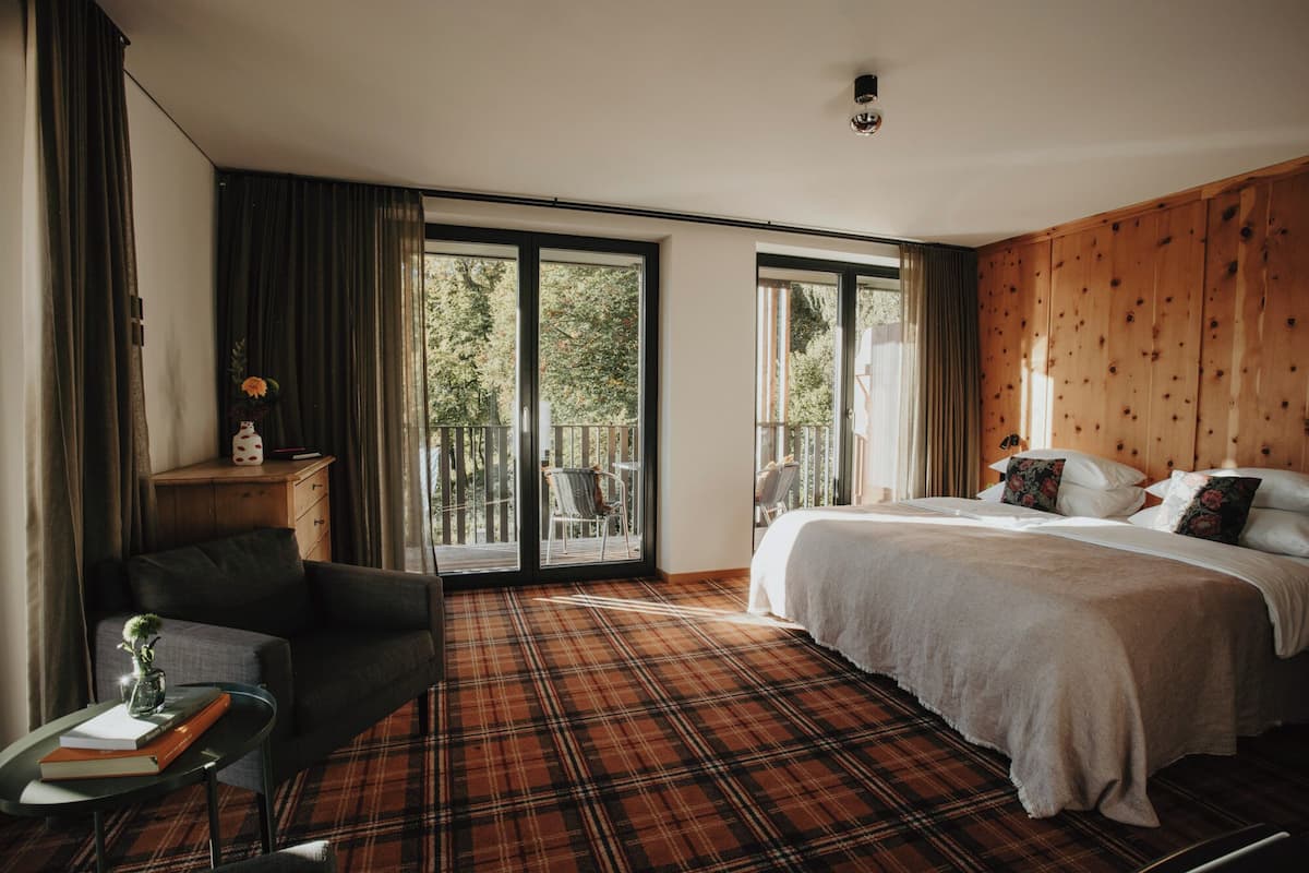 Une chambre à coucher avec un lit, une chaise, une petite table avec des livres et une vue dégagée grâce à de grandes fenêtres.