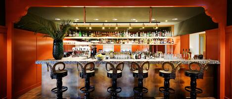 Una zona de bar con un mostrador central, varios taburetes y una variedad de botellas y vasos.