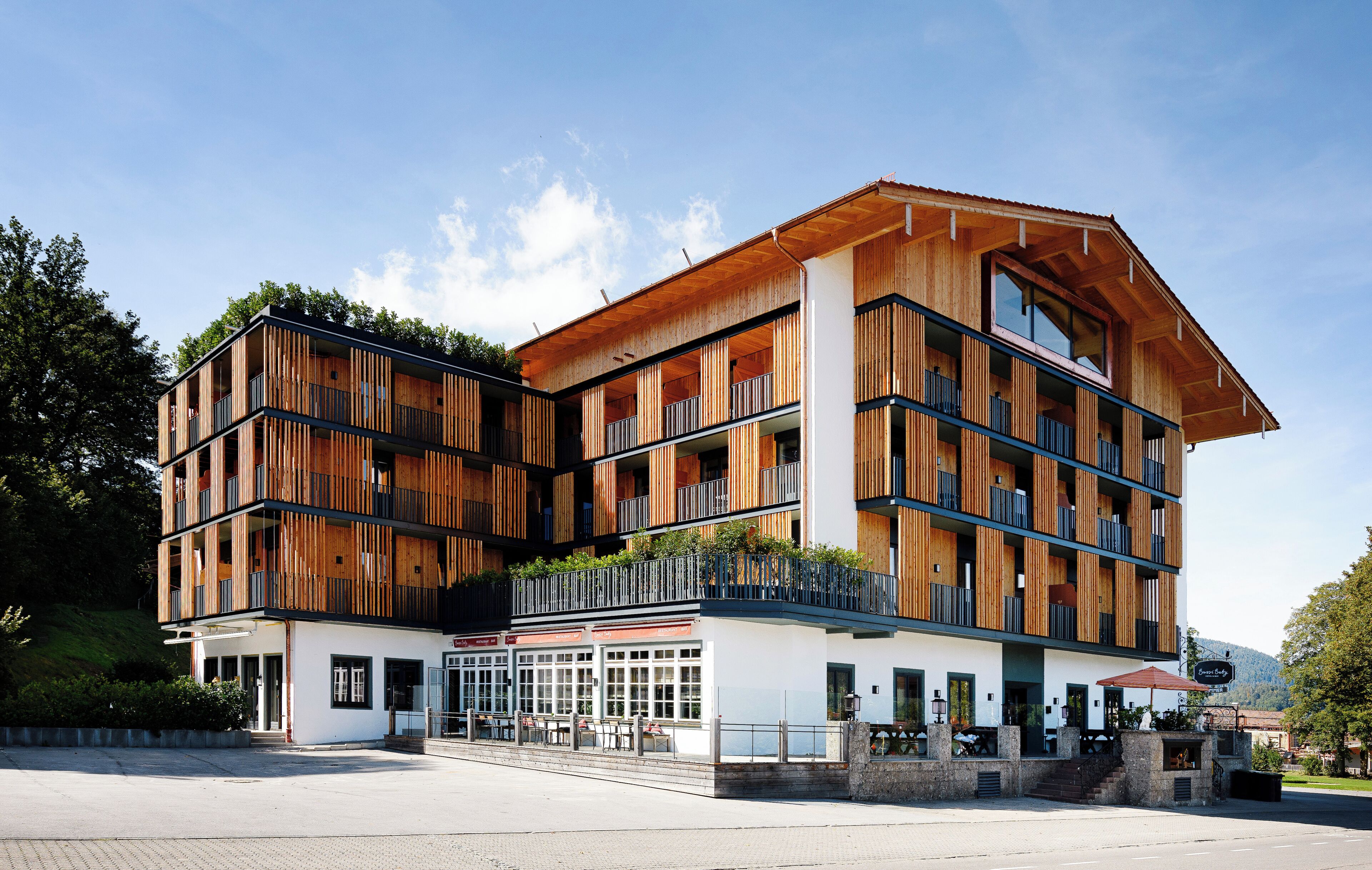 Ein mehrstöckiges Gebäude mit Holzfassade, Balkonen und einem Eingangsbereich mit Außenbestuhlung.