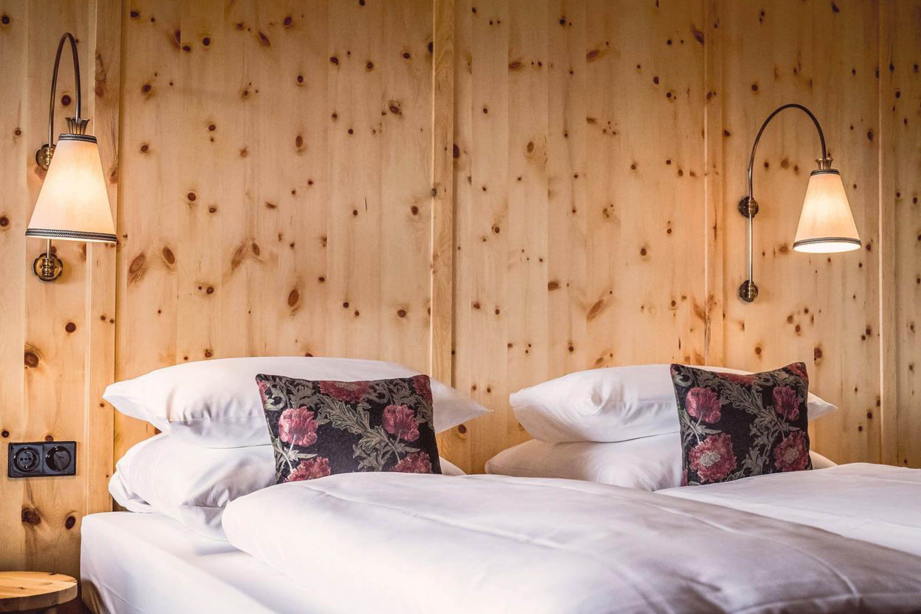 A wooden-paneled room with two beds, each with white bedding and floral-patterned pillows. There are two wall-mounted lamps with a classic design.