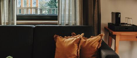 Una sala de estar moderna con un sofá negro, dos almohadas marrones, una mesita con un florero y libros, y un televisor sobre un mueble de madera.