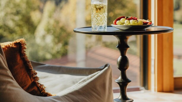 Une table avec un verre et une assiette de fruits, une chaise rembourrée et, par la fenêtre, une vue sur un paysage montagneux.