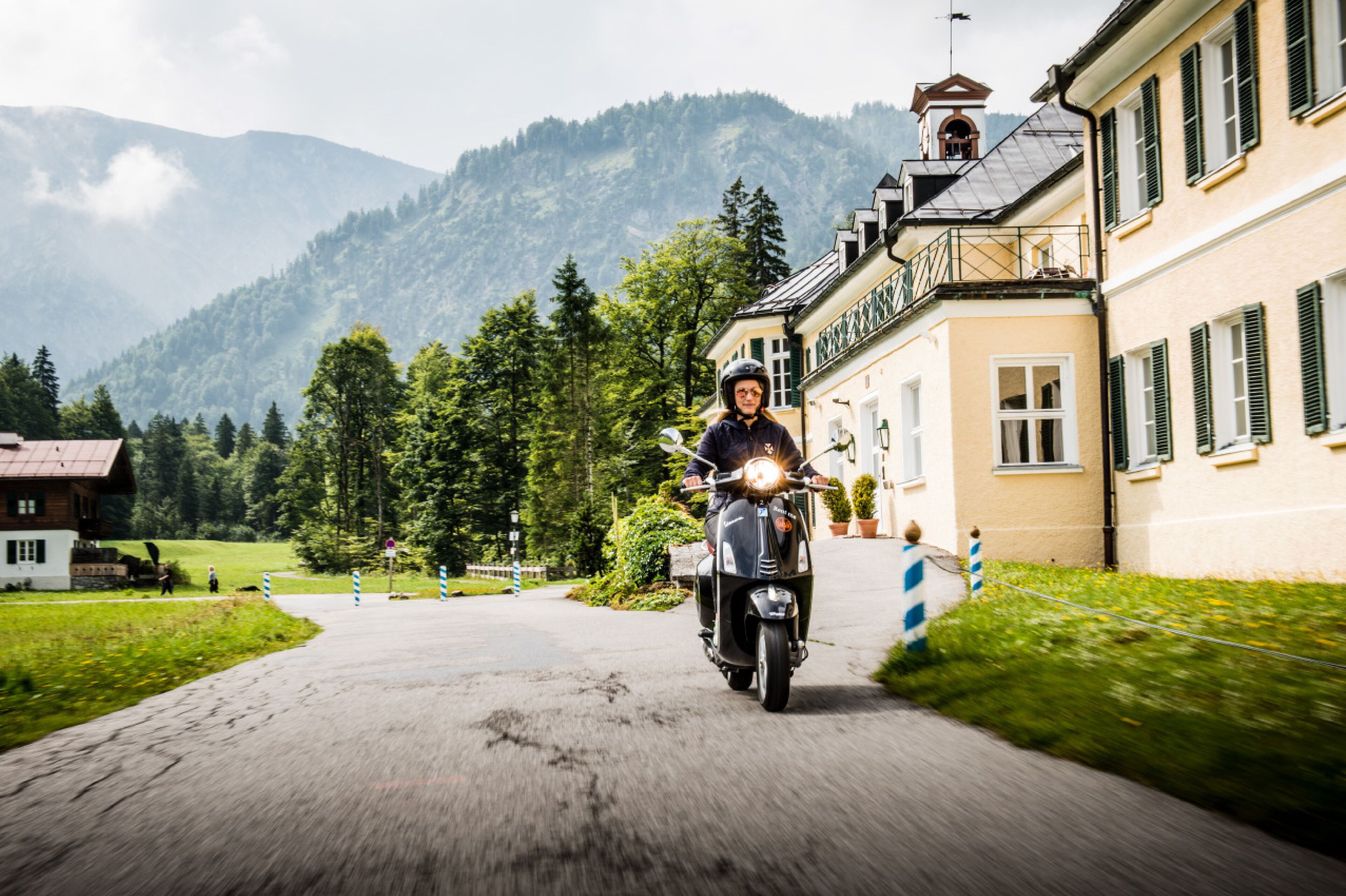 A person riding a scooter on a paved road with a building on the right and mountains in the background.