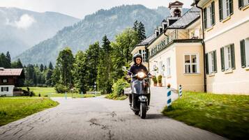 A person riding a scooter on a paved road with a building on the right and mountains in the background.