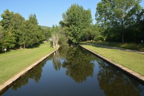 A canal with a narrow path on one side and a grassy area on the other, surrounded by trees.
