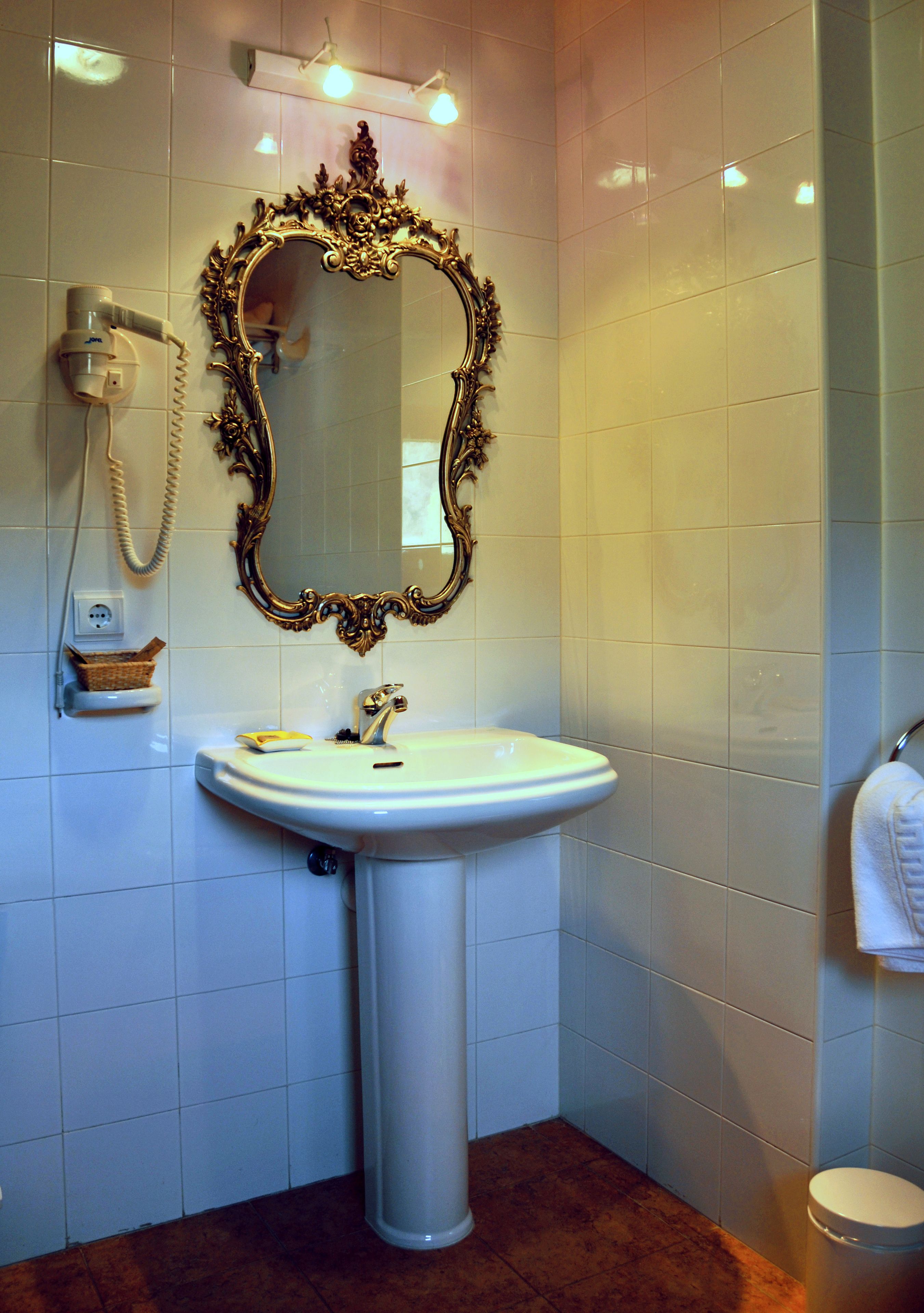 Un baño con lavamanos pedestal, un espejo grande y ornamentado, un secador de pelo en la pared y un perchero para toallas.