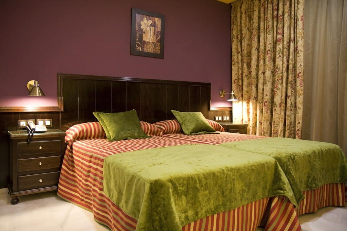 Una cama doble con cubrecamas verdes, una mesita de noche de madera con lámpara, teléfono y un cuadro en la pared.