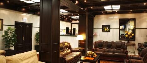 Un vestíbulo de hotel con un sofá beige, un sofá de cuero marrón y un sillón de cuero marrón. Hay una mesa de centro de madera con un jarrón de flores. El vestíbulo tiene pisos de madera y un tapete a cuadros.