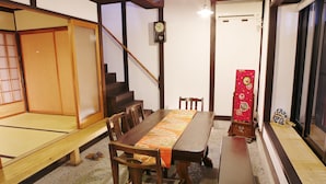 Una habitación japonesa tradicional con suelo de tatami, una mesa de madera para comer con camino de mesa estampado, una silla de madera, un reloj en la pared y una puerta corredera.