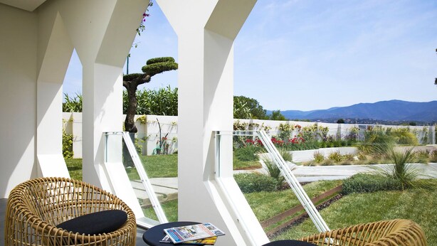 Un balcon agrémenté de fauteuils et d’une table en osier, offrant une vue sur un jardin aménagé et les montagnes.