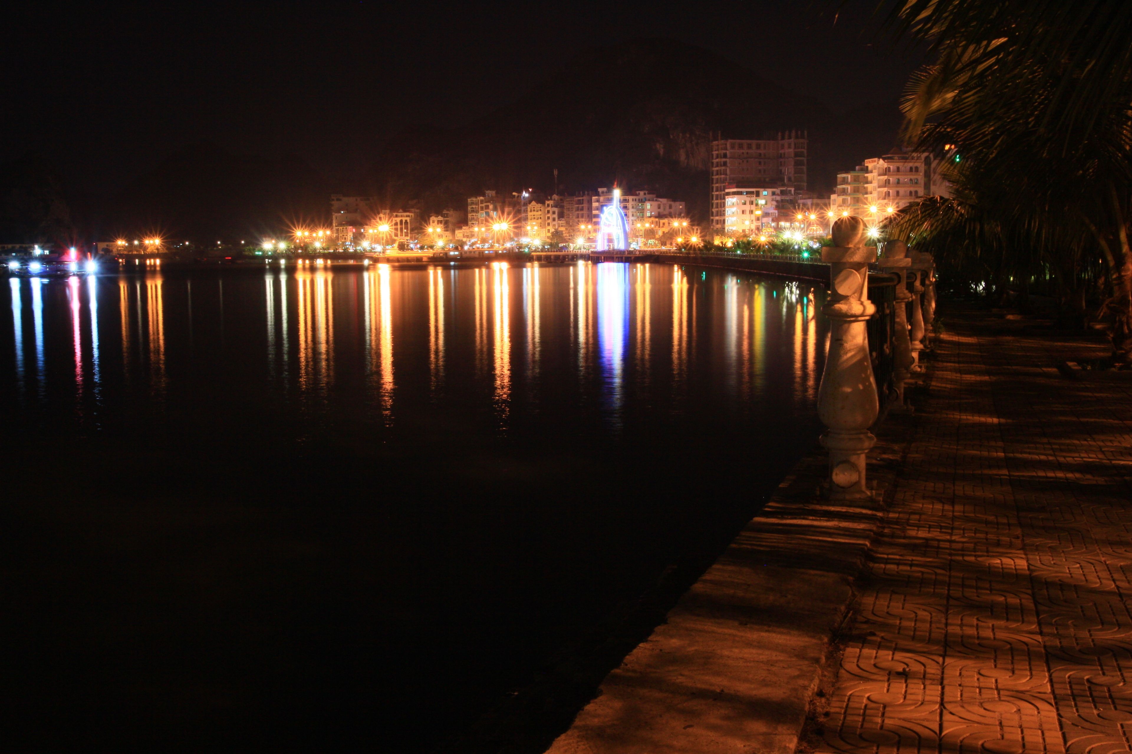 Vista noturna de um horizonte urbano à beira-mar, com edifícios iluminados refletindo na água.