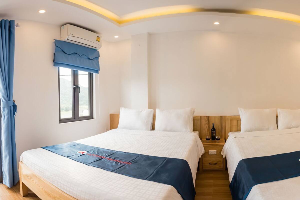 Ein Hotelzimmer mit zwei Betten, einem Holzschrank, einem Fenster mit Vorhängen, einer Klimaanlage und einem Nachttisch mit Flasche und Gläsern.