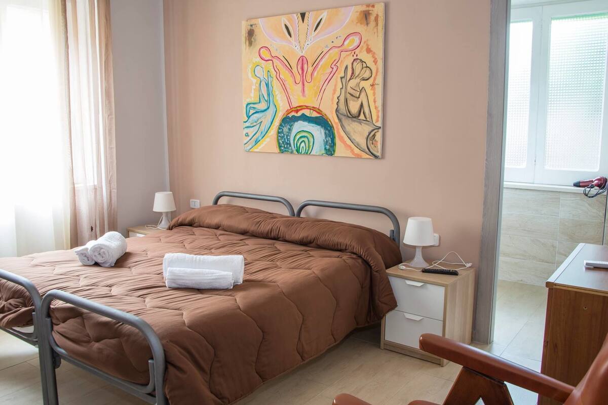 A bedroom with a brown bed, two bedside tables with lamps, a painting on the wall, and a view of the outside through a window.