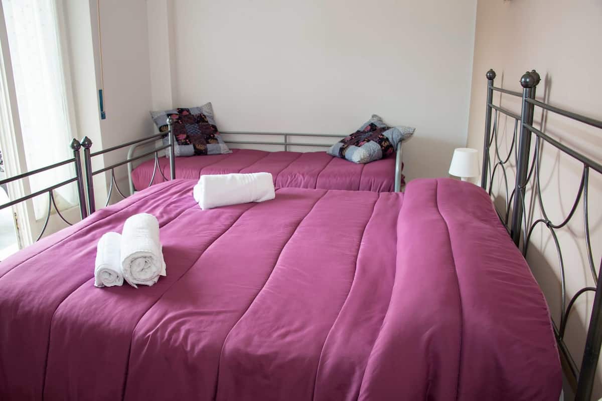 A double bed with a pink bedspread and two pillows, a white lamp on a bedside table, and a window with curtains.