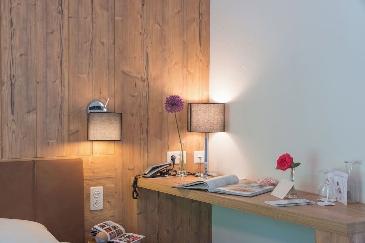 A hotel room with a wooden headboard, a bedside table with a lamp, a phone, a vase with a flower, and a magazine.