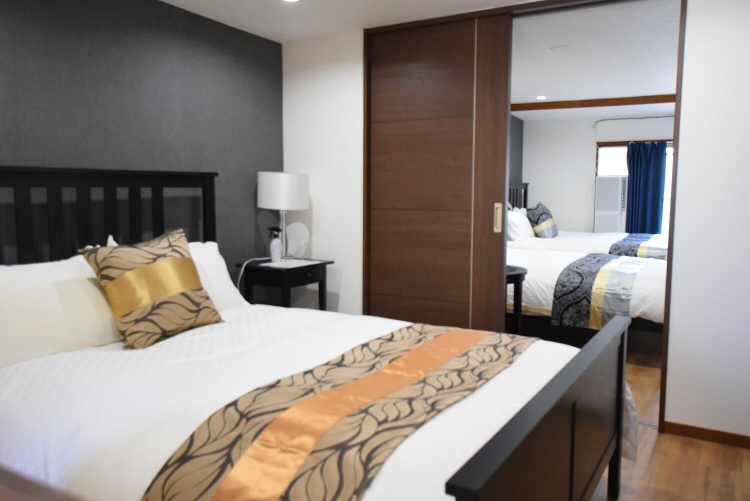 A hotel room with a large bed, a nightstand with a lamp, a mirror, and a view of another room.