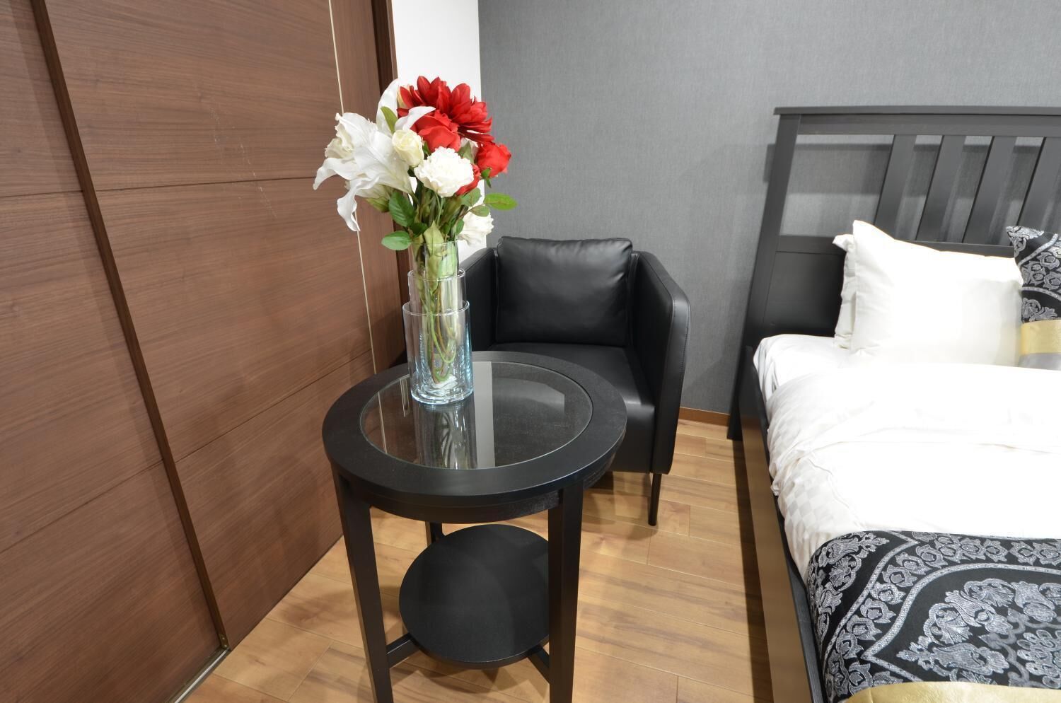 A modern hotel room with a round glass table, a black leather chair, a bed with patterned bedding, and a wooden wardrobe.