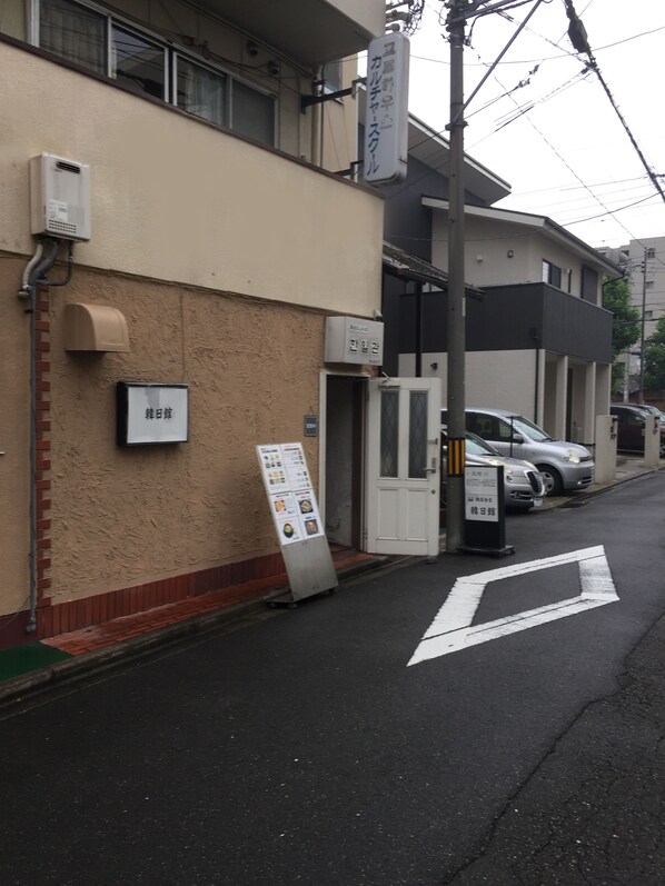 A street view with a building that has a beige exterior, a mailbox, and a signboard. There are parked cars and a pedestrian crossing. - Kyoto House (Kyoto)
