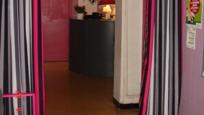 A hallway with pink walls, a kitchen area in the background, and a door with striped curtains. - Hotel La Marmotte (Niort)