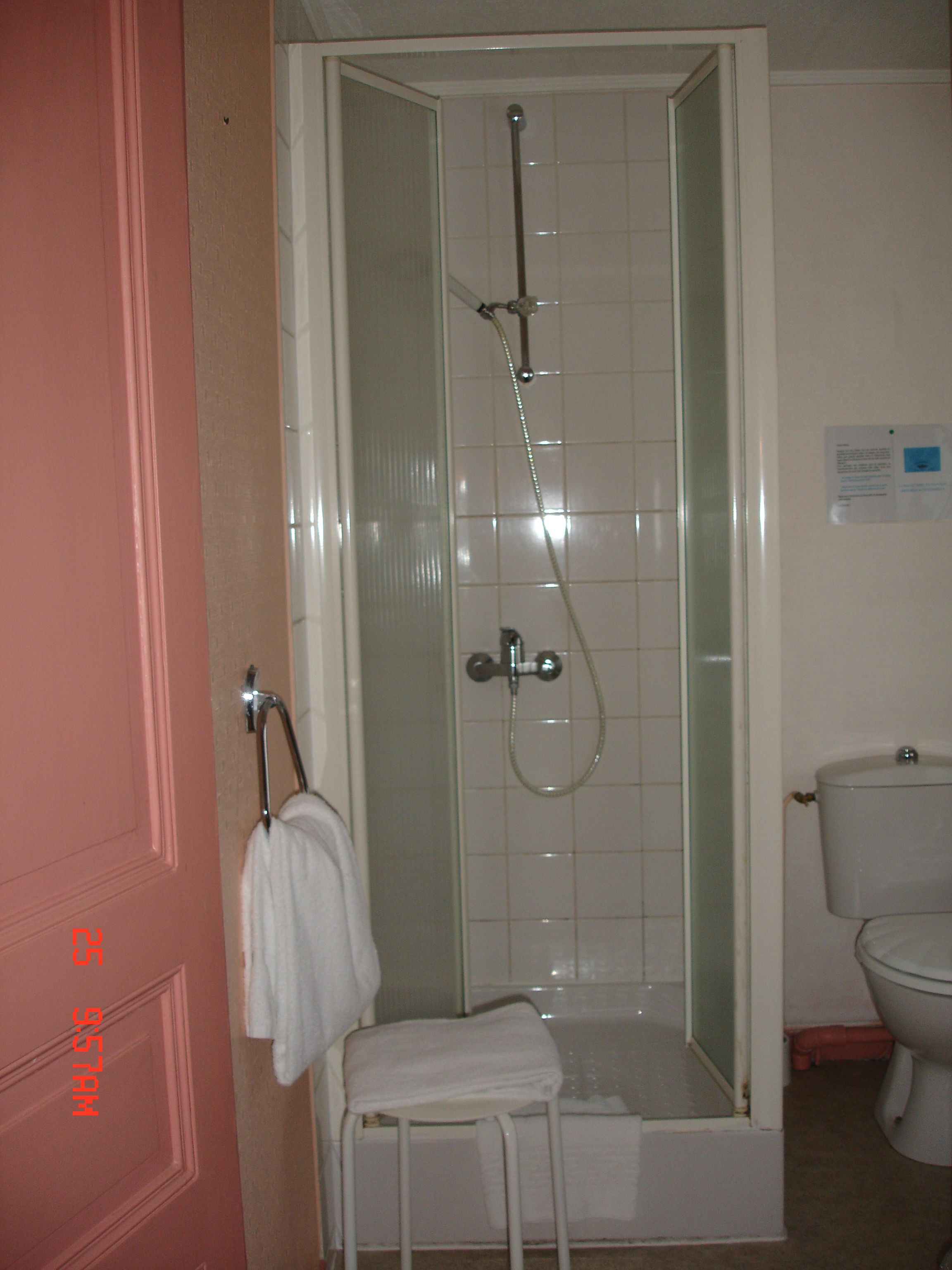 Une douche avec une porte en verre, une serviette blanche sur un porte-serviettes et des toilettes dans la pièce adjacente.