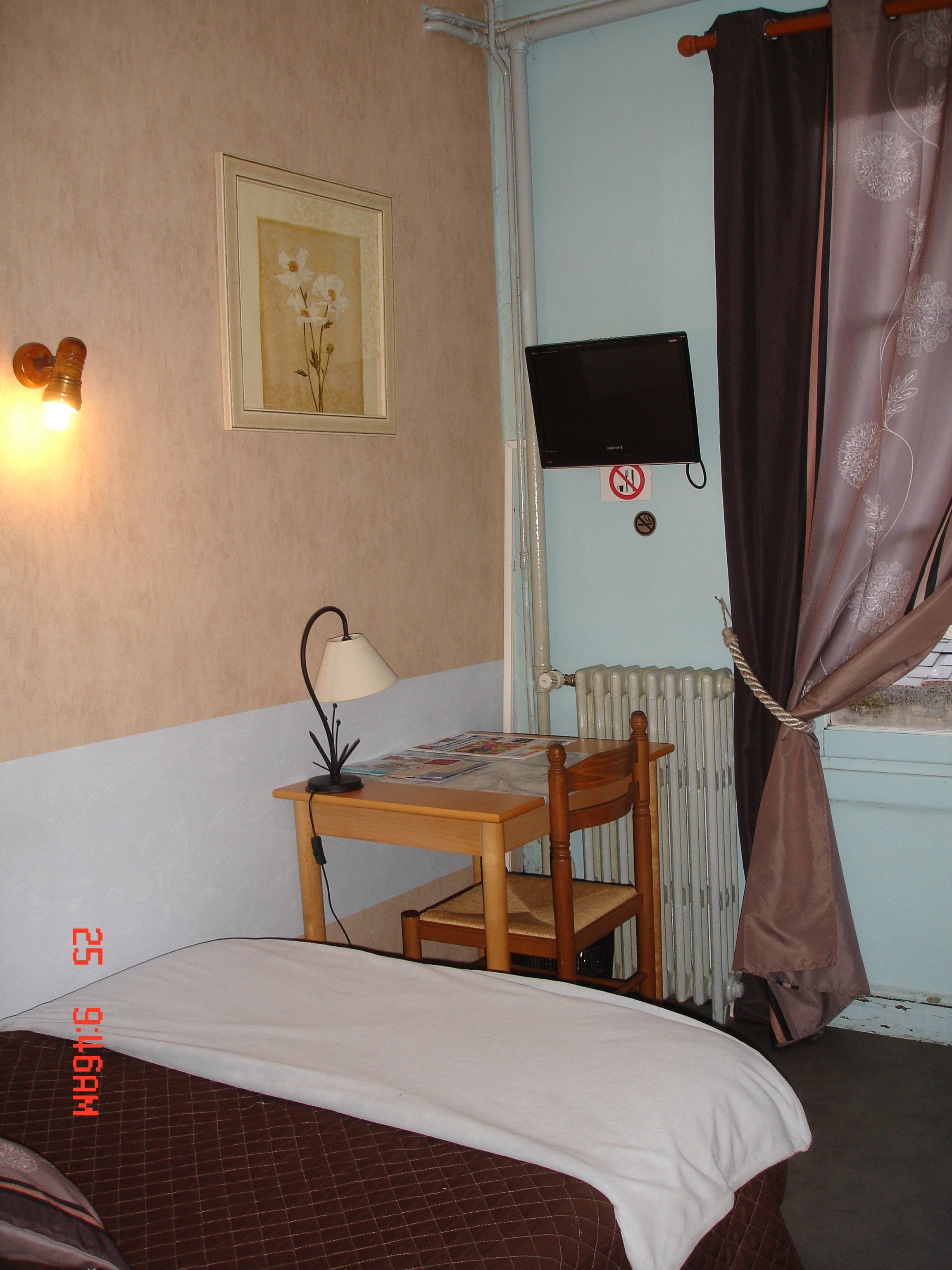 Een kleine kamer met een bed, een bureau met een lamp, een stoel, een televisie en een radiator.