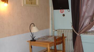 Een kleine kamer met een bed, een bureau met een lamp, een stoel, een televisie en een radiator.