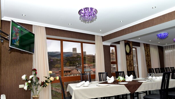 A dining room with a table set for six, large windows with a view of a castle, a chandelier, and a flat-screen TV mounted on the wall. - Grand Palace (Akhaltsikhe)