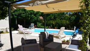 Un espace extérieur aménagé avec une zone salon couverte, des meubles en osier, une piscine et une végétation luxuriante en arrière-plan.