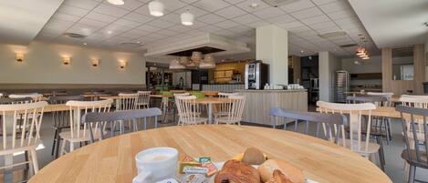 A spacious cafeteria with multiple tables and chairs, a variety of food items including croissants and yogurt, and a coffee station.