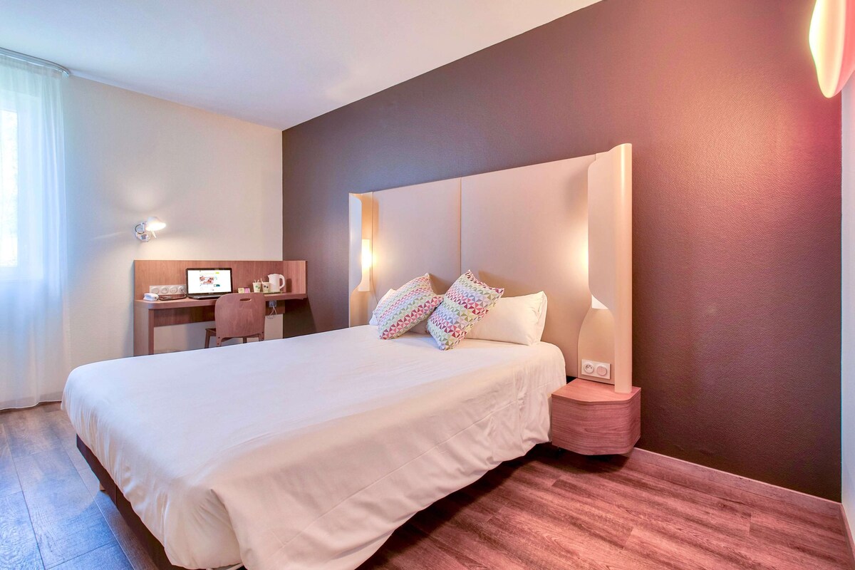 A hotel room with a large bed, a desk with a computer, and a nightstand with a lamp.