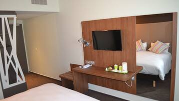 A hotel room with a bed, a TV mounted on a wooden panel, a small desk with a telephone and a cup, and a mirror reflecting the bed and pillows.