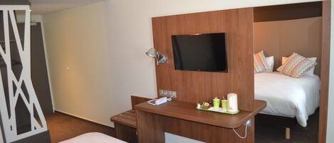 Une chambre d’hôtel avec un lit, un téléviseur fixé sur un panneau en bois, un petit bureau avec un téléphone et une tasse, et un miroir qui reflète le lit et les oreillers.