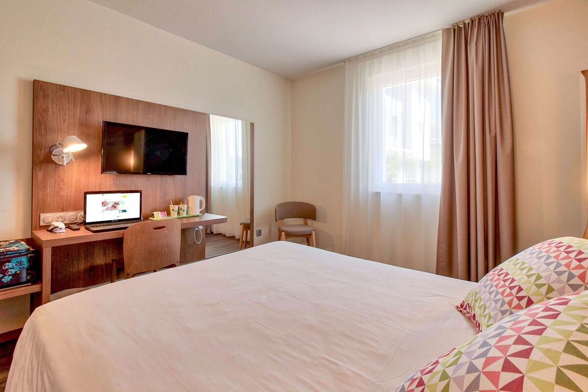 A hotel room with a large bed, a desk with a computer, a TV mounted on the wall, and a window with curtains.