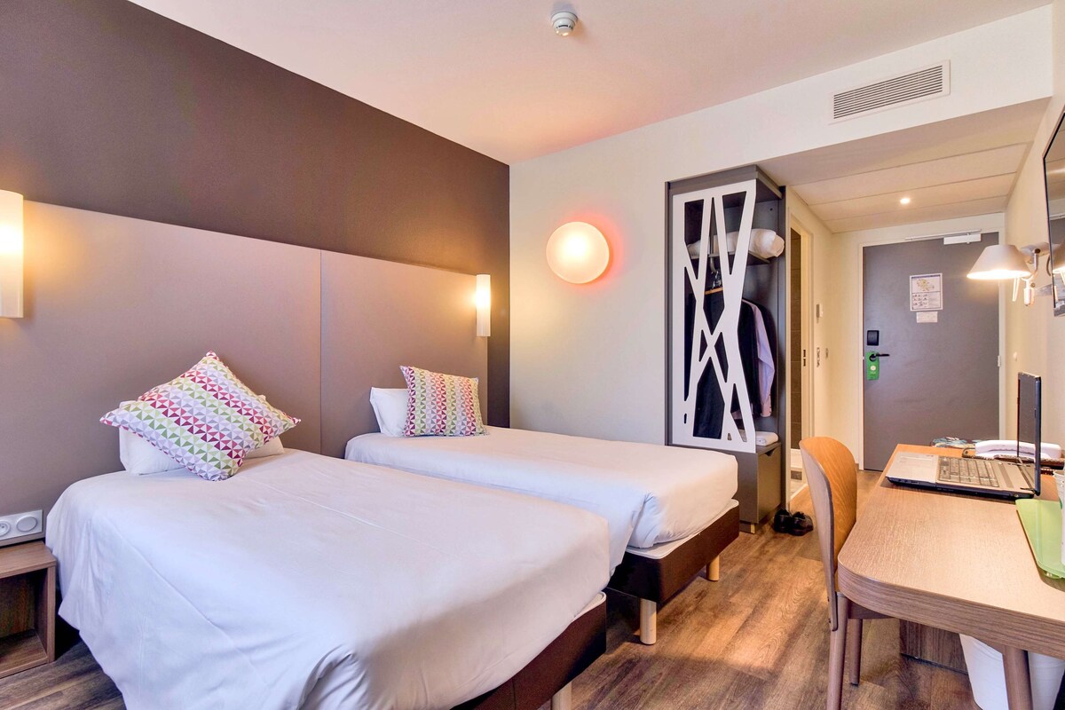 A hotel room with two beds, a desk with a laptop, a chair, and a mirror.