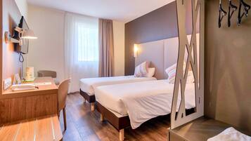 A hotel room with two beds, a desk, and a chair. There is a window with curtains, a wall-mounted TV, and a coat rack.