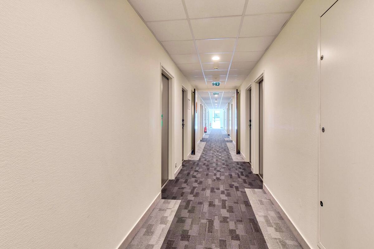 Un long couloir étroit, dont le sol est recouvert de moquette, et de part et d’autre duquel se trouvent des portes menant à différentes pièces.